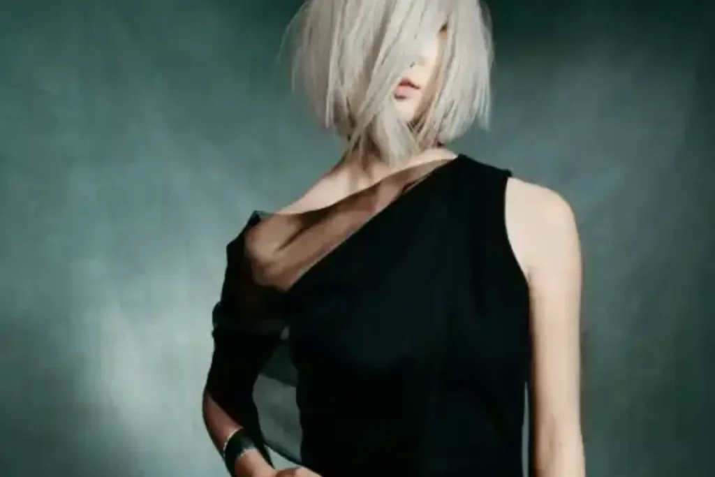 A person with light blonde, chin-length hair styled asymmetrically, enhanced with Aveda Demi Hair Color for healthy hair, wears a black one-shoulder dress against a dark, blurred background.
