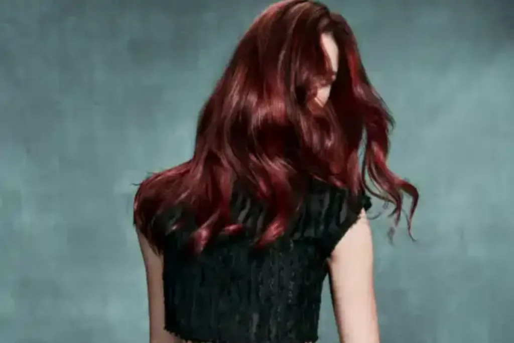 A person with long, healthy, wavy reddish-brown hair poses with their back mostly to the camera, wearing a sleeveless black top against a plain gray background.