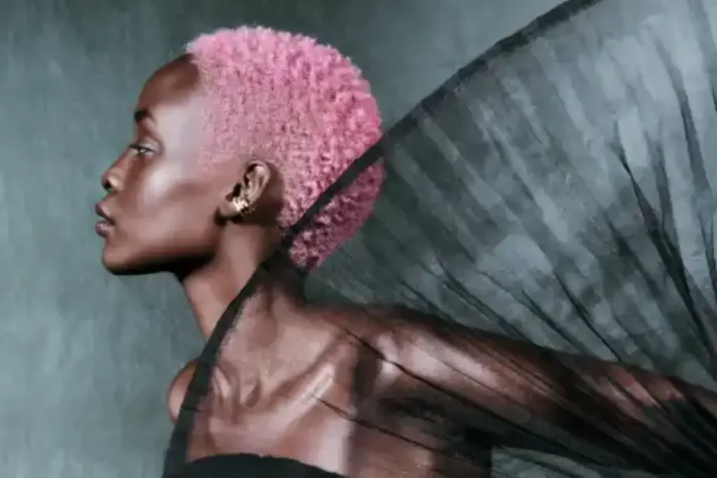 Person with short, curly pink hair showcases radiant hair in profile against a neutral background, with sheer black fabric draped dramatically behind them—highlighting the vibrance of Demi Hair Color.
